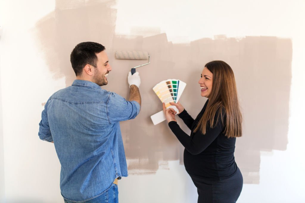Young pregnant woman choosing color from palette for walls while husband painting with roller.